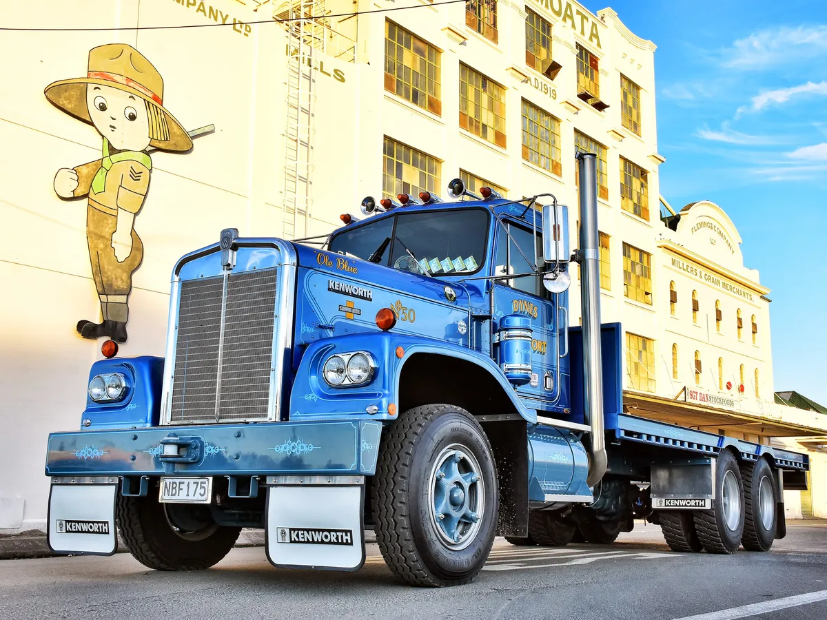 A blue Kenworth W-model bonneted truck parked outside an old cream brick mill building