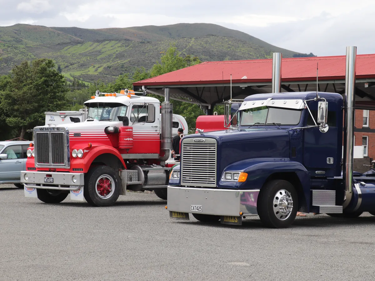 A red bonneted Kenworth and a navy blue Kenworth parked under a red roofed shelter with hills behind