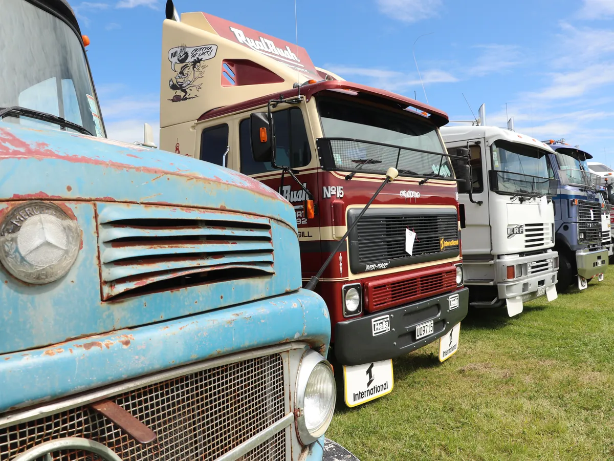 A row of trucks on grass at a show, an unrestored pale blue Mercedes bonnet closest to the camera