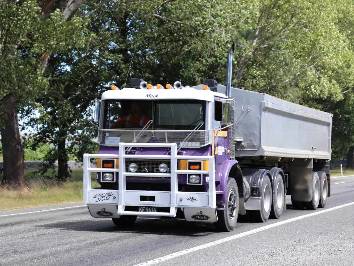 A purple and white Mack pulling a loaded tipping trailer along a tree lined road