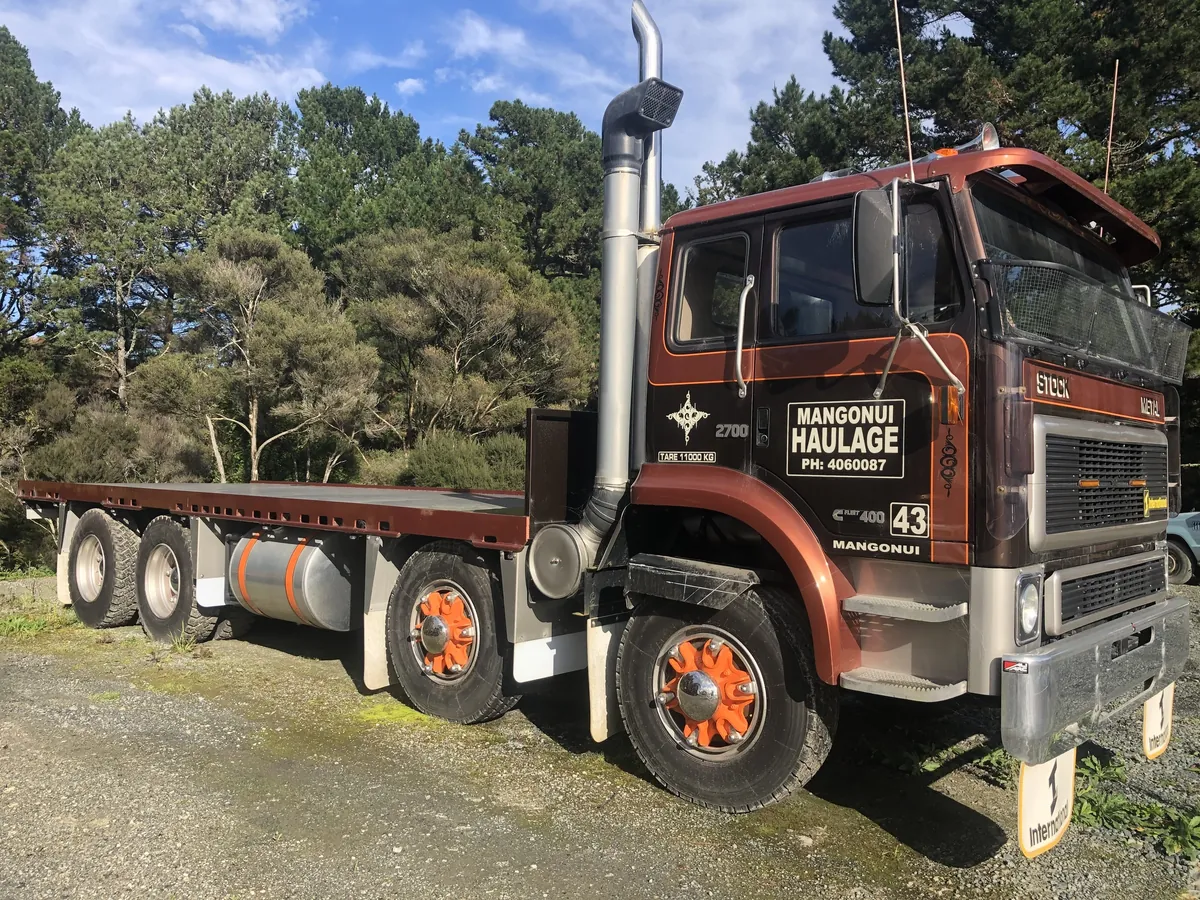 A brown International flatdeck signwritten Mangonui Haulage, parked on gravel beside pines