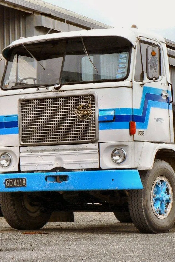 A 1972 Volvo cab-over truck in white with a blue stripe, parked beside a shed