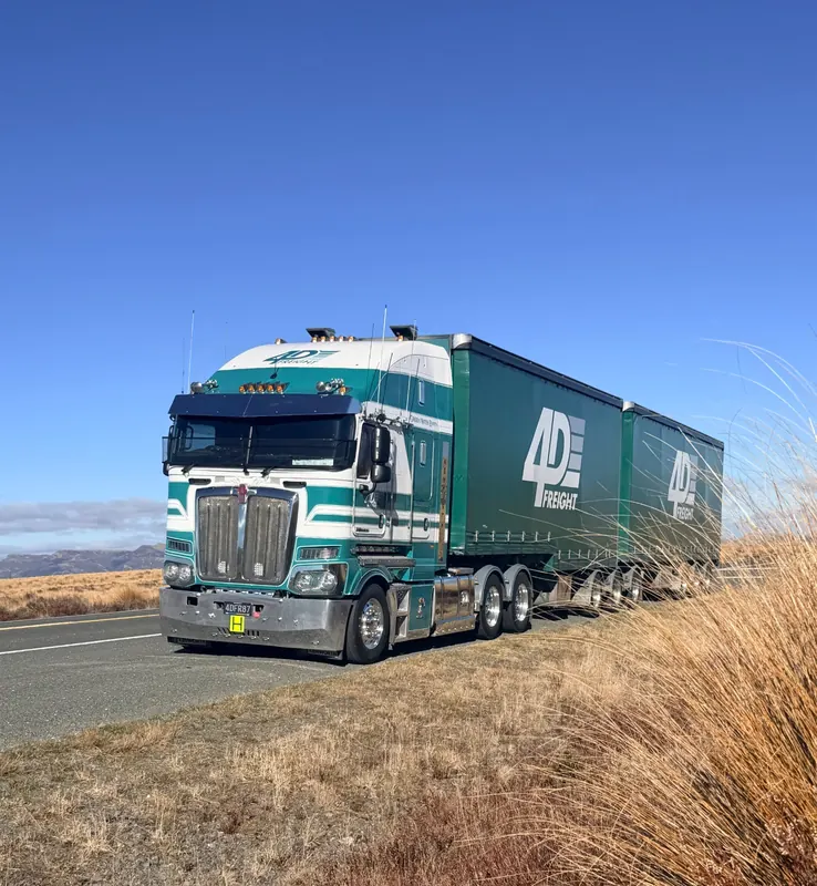 A green and white Kenworth K220 curtainsider unit beside tussock